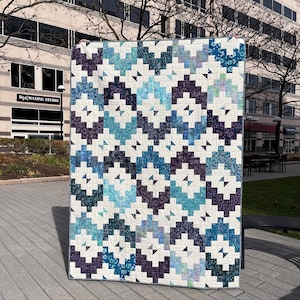 May include: A quilt with a geometric pattern in shades of blue, purple, and white. The quilt is made up of squares and triangles, and the pattern is repeated throughout the quilt.