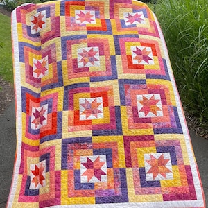 May include: A colorful quilt with a patchwork design featuring a variety of shades of pink, yellow, orange, and purple. The quilt is made up of squares with a star pattern in the center of each square.