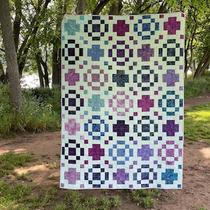 Może przedstawiać: Duża, ręcznie robiona kołdra z powtarzającym się wzorem kwadratów, krzyży i kół w odcieniach fioletu, niebieskiego i turkusowego na kremowym tle. Kołdra jest prezentowana na zewnątrz, na tle drzew i zieleni.