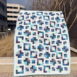 May include: A large, handmade quilt with a white background and a repeating pattern of blue, teal, and purple crosses and squares. The quilt has a light blue border and is displayed outdoors.