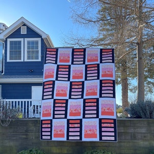 May include: A pink and white quilt with black squares and a floral pattern. The quilt is hanging on a clothesline in front of a blue house.