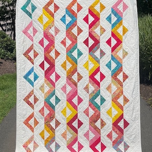 Pode incluir: Um edredom branco com um padrão geométrico de triângulos coloridos. Os triângulos são dispostos em filas verticais, cada fila com uma combinação de cores diferente. As cores incluem vermelho, rosa, amarelo, azul e verde.