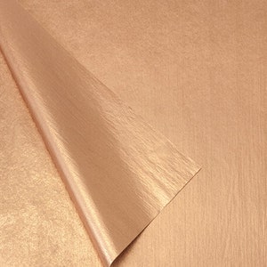 May include: A sheet of rose gold metallic paper with a textured surface. The corner of the paper is folded over, revealing the underside. The paper has a subtle sheen and a warm, inviting color.