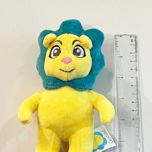 May include: A yellow plush lion with a teal mane and a bow tie. The lion is standing on a purple surface and has a tag that says "CREATIONS".
