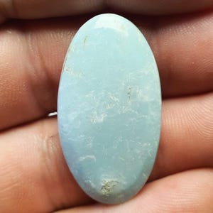 May include: A light blue oval cabochon gemstone with a smooth, polished surface. The stone has a cloudy, marbled appearance with subtle white and gray inclusions.