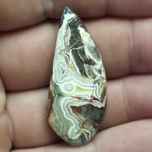 Peut inclure: Un cabochon d'agate en forme de larme avec un motif tourbillonnant de blanc, brun et gris. La pierre a une finition naturelle et polie.