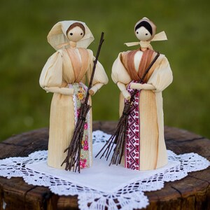 Pode incluir: Duas bonecas artesanais de palha de milho com cabeças de contas de madeira lisa e vestidos longos em tons bege claro. Cada figura usa um lenço na cabeça e segura um pequeno feixe de galhos castanhos escuros. Uma boneca tem um avental floral.