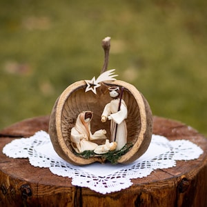 Può includere: Un presepe realizzato con foglie di mais essiccate all'interno di un guscio di noce marrone svuotato. Le figure includono una persona inginocchiata, una in piedi con un bastone e un piccolo neonato, con aureole semplici e una stella.