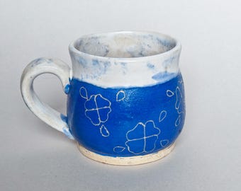May include: A blue ceramic mug with a white rim and white floral designs. The mug has a rounded body and a handle.
