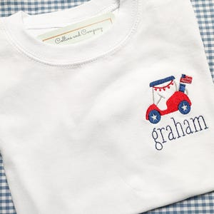 May include: White crew neck t-shirt with a red and blue embroidered design of a vehicle with an American flag and the name "graham" in blue. The shirt has a tag that reads "Collins and Company".