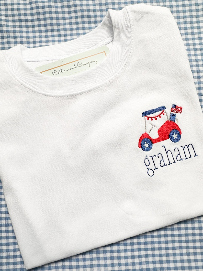 May include: White crew neck t-shirt with a red and blue embroidered design of a vehicle with an American flag and the name "graham" in blue. The shirt has a tag that reads "Collins and Company".