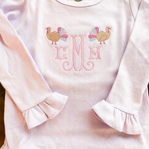 May include: A light purple long-sleeved shirt with ruffled cuffs and hem. The shirt features an embroidered design of two turkeys and a monogram in pink thread. A small tag is visible near the neckline.