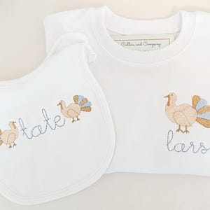 May include: A white baby bib and a white t-shirt with embroidered turkeys and the names "Tate" and "Lars" in blue thread.