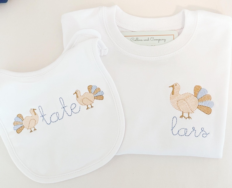 May include: A white baby bib and a white t-shirt with embroidered turkeys and the names "Tate" and "Lars" in blue thread.