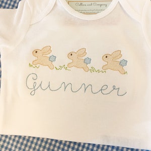 May include: White baby onesie with three embroidered brown bunnies hopping in a line. The onesie has the name "Gunner" embroidered in blue thread below the bunnies.
