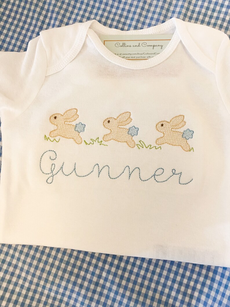 May include: White baby onesie with three embroidered brown bunnies hopping in a line. The onesie has the name "Gunner" embroidered in blue thread below the bunnies.