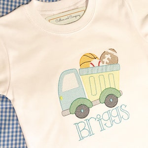May include: A white t-shirt with a blue and green truck carrying a basketball, baseball, and football. The name "Briggs" is embroidered below the truck.