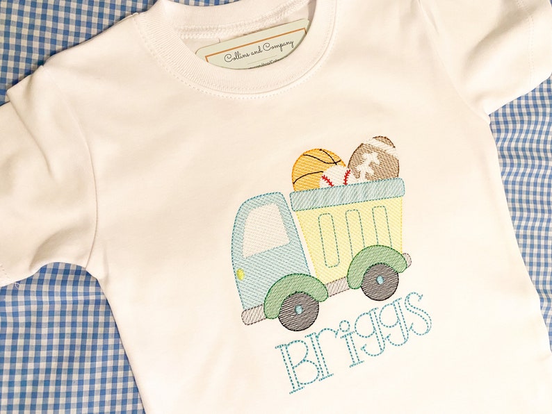 May include: A white t-shirt with a blue and green truck carrying a basketball, baseball, and football. The name "Briggs" is embroidered below the truck.