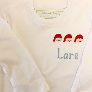 Op de afbeelding: Een wit longsleeve shirt met een blauwe geborduurde naam "Lars" en drie rode en witte kerstman figuren.