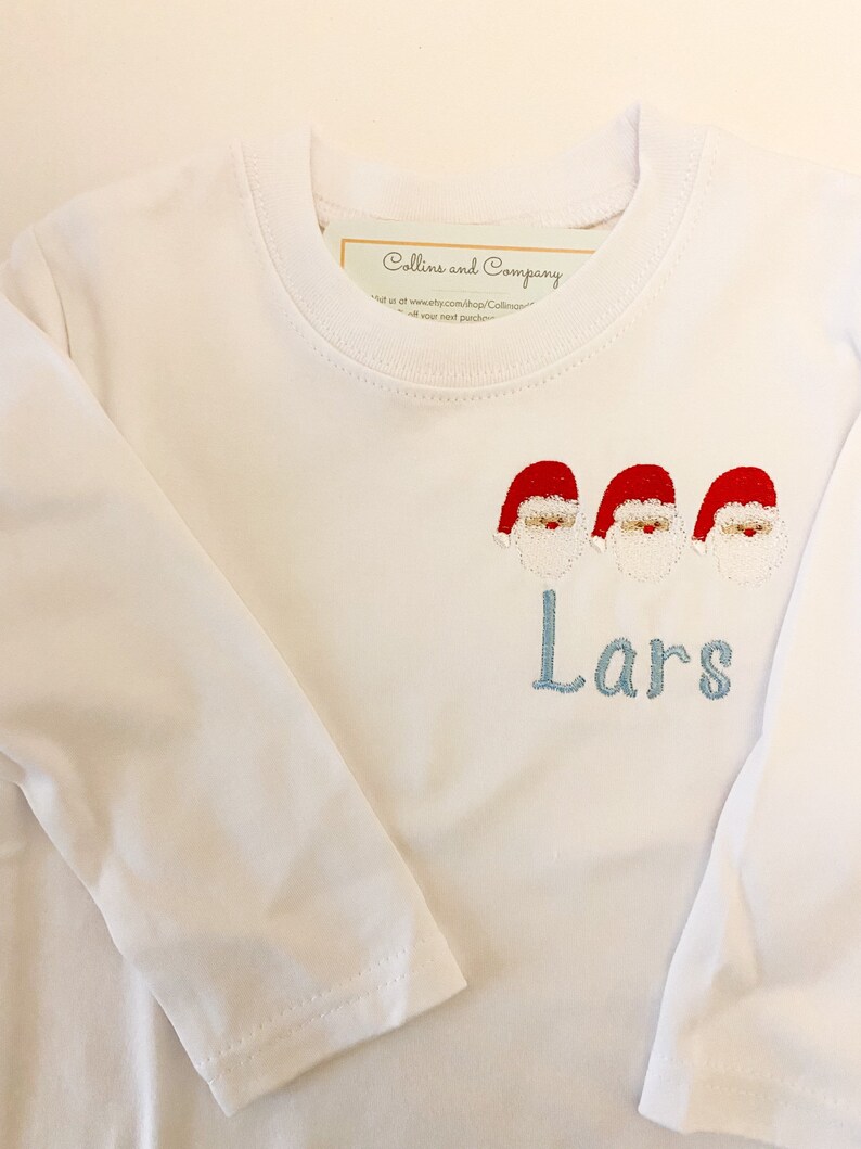 Op de afbeelding: Een wit longsleeve shirt met een blauwe geborduurde naam "Lars" en drie rode en witte kerstman figuren.