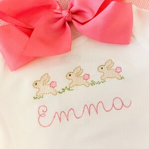 May include: White baby onesie with three embroidered bunnies and the name "Emma" in pink thread. A pink bow is on top of the onesie.