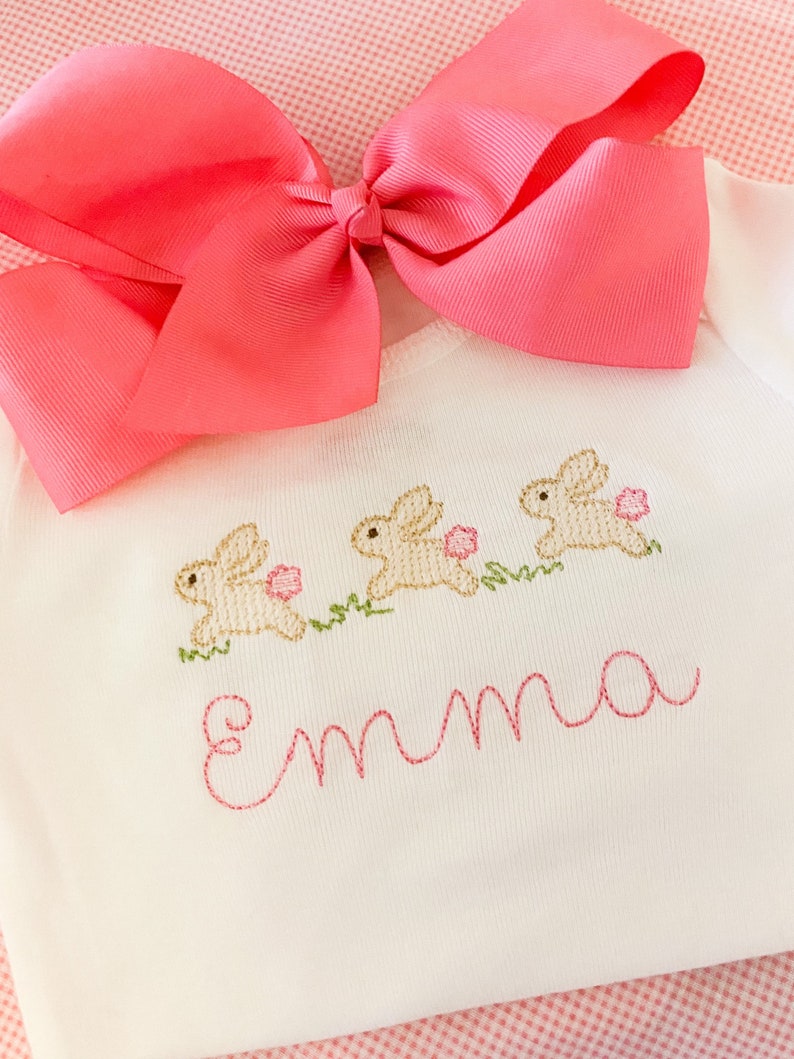 May include: White baby onesie with three embroidered bunnies and the name "Emma" in pink thread. A pink bow is on top of the onesie.