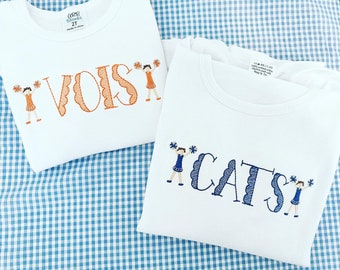 May include: Two white t-shirts with embroidered cheerleaders and the words "VOIS" and "CATS" in orange and blue.
