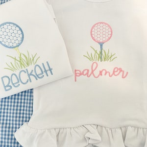 May include: Two white shirts with embroidered golf ball designs and names. The shirt on the left has a blue golf ball and the name "Beckett" embroidered in blue. The shirt on the right has a pink golf ball and the name "palmer" embroidered in pink.