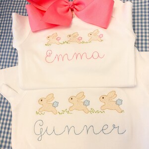 May include: Two white onesies with embroidered bunnies and names. The top onesie has pink bunnies and the name "Emma" embroidered in pink. The bottom onesie has blue bunnies and the name "Summer" embroidered in blue.