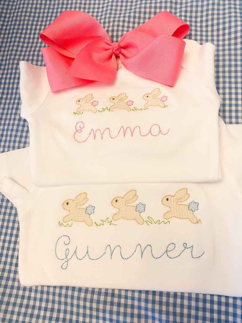 May include: Two white onesies with embroidered bunnies and names. The top onesie has pink bunnies and the name "Emma" embroidered in pink. The bottom onesie has blue bunnies and the name "Summer" embroidered in blue.