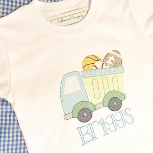 May include: A white t-shirt with a blue truck carrying a basketball, baseball, and football. The name "Briggs" is embroidered below the truck in blue thread.