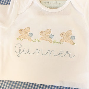 May include: White baby onesie with three embroidered brown bunnies hopping with blue flowers. The onesie has the name "Gunner" embroidered in blue thread.