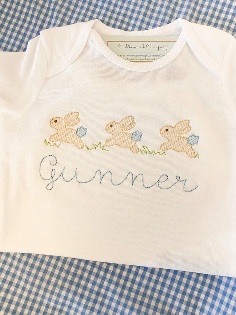 May include: White baby onesie with three embroidered brown bunnies hopping with blue flowers. The onesie has the name "Gunner" embroidered in blue thread.