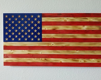 Op de afbeelding: Een Amerikaanse vlag van hout met rode, witte en blauwe strepen. De sterren zijn goudkleurig en zijn verhoogd op het blauwe veld. De vlag is gemaakt van gerecycled hout en heeft een rustieke uitstraling.