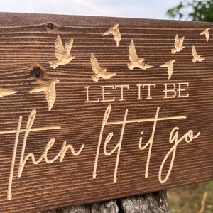 May include: A wooden sign with a dark brown stain. The sign has a carved design of birds flying away. The text on the sign reads "Let it be then let it go."