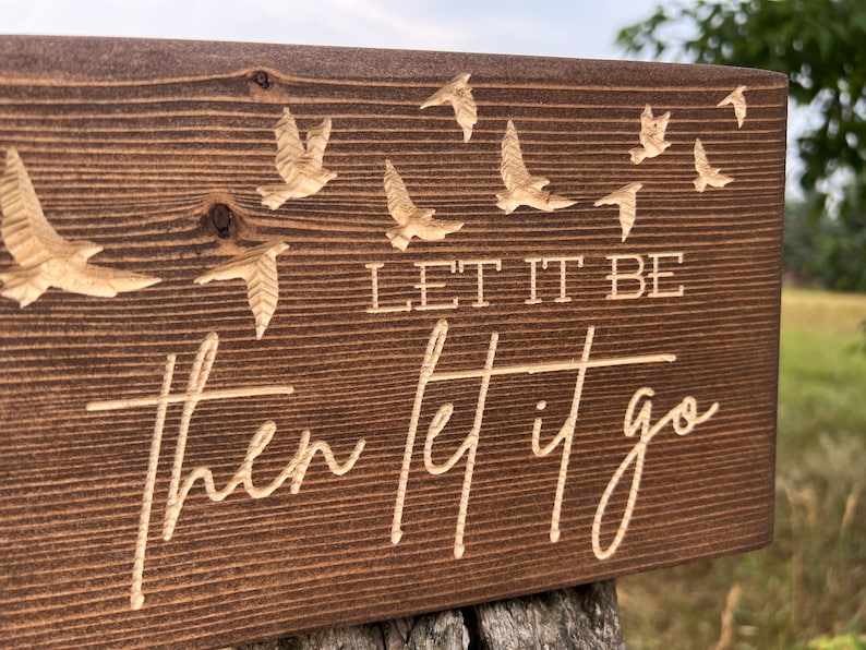 May include: A wooden sign with a dark brown stain. The sign has a carved design of birds flying away. The text on the sign reads "Let it be then let it go."
