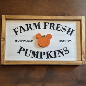 Könnte beinhalten: Rechteckiges Holzschild mit den schwarzen Worten "FARM FRESH PUMPKINS". In der Mitte befindet sich ein orangefarbener Kürbis mit Mickey Mouse Ohren. Das Schild trägt auch die Aufschrift "HAND PICKED" und "SINCE 1955".
