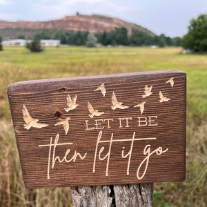 May include: A wooden sign with a dark brown stain. The sign has a white inscription that reads "Let it be then let it go". The inscription is surrounded by a pattern of flying birds.
