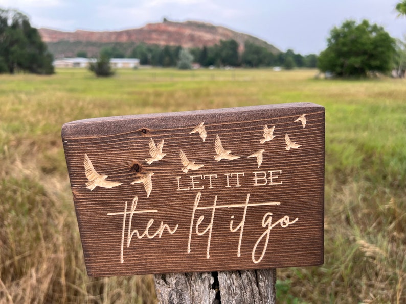 May include: A wooden sign with a dark brown stain. The sign has a white inscription that reads "Let it be then let it go". The inscription is surrounded by a pattern of flying birds.
