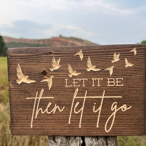 May include: A wooden sign with a brown stain and a light brown engraved design. The sign features a quote that reads "Let it be then let it go" with a series of birds flying above the text.