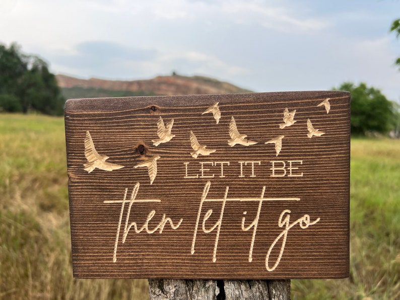 May include: A wooden sign with a brown stain and a light brown engraved design. The sign features a quote that reads "Let it be then let it go" with a series of birds flying above the text.