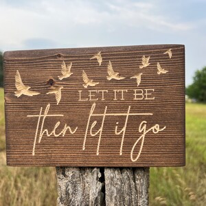 May include: A wooden sign with the text "Let it be then let it go" engraved in a light brown colour. The sign is decorated with a flock of birds flying in the background.