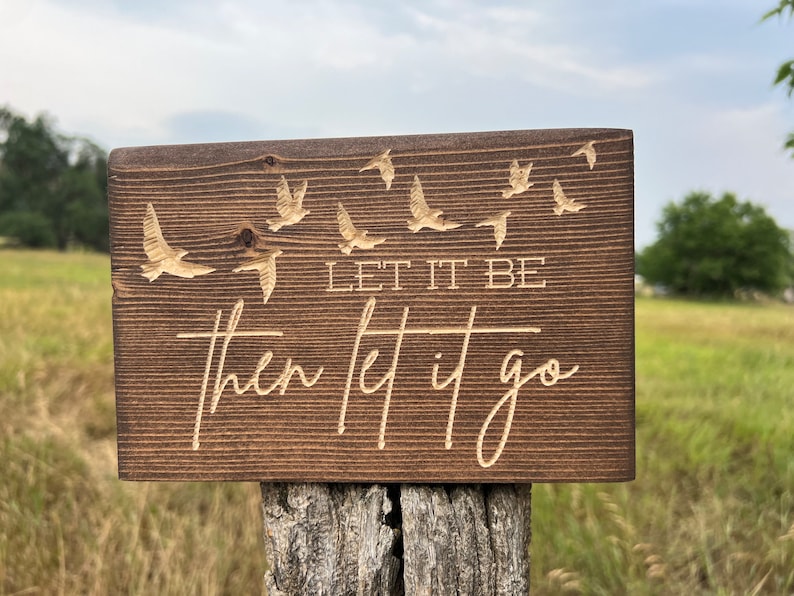 May include: A wooden sign with the text "Let it be then let it go" engraved in a light brown colour. The sign is decorated with a flock of birds flying in the background.