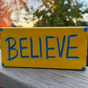 May include: A rectangular yellow wooden block with the word BELIEVE carved into the front face. The letters are painted in a bright blue color, and small blue accents are visible on each of the four corners of the block's front surface.