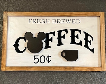 May include: A wooden sign with a brown frame, white background, and black lettering. The sign reads "FRESH BREWED COFFEE" with a Mickey Mouse head and a coffee cup silhouette. The price is listed as "50¢".