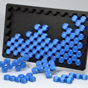 Op de afbeelding: Een blauwe zeshoekige puzzel met stukken die rond een zwart frame zijn verspreid. De puzzel is gemaakt van plastic en heeft een honingraatpatroon.