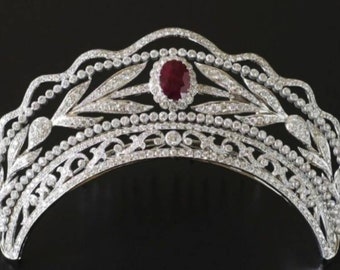 May include: A silver tiara with a large red ruby gemstone in the center, surrounded by smaller diamonds. The tiara is decorated with a delicate floral design.