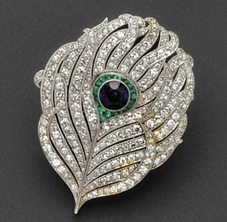 May include: A silver peacock feather brooch with a blue sapphire and emerald accents. The feather is set with diamonds and has a detailed design.