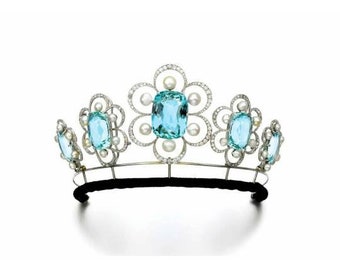 Puede incluir: Una tiara de plata con una piedra preciosa de aguamarina central rodeada de piedras preciosas de aguamarina más pequeñas y perlas. La tiara está colocada sobre una base de terciopelo negro.