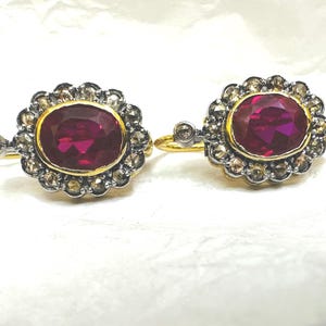 May include: A pair of gold earrings with a ruby gemstone surrounded by a halo of smaller diamonds. The earrings have a silver-toned hook closure.